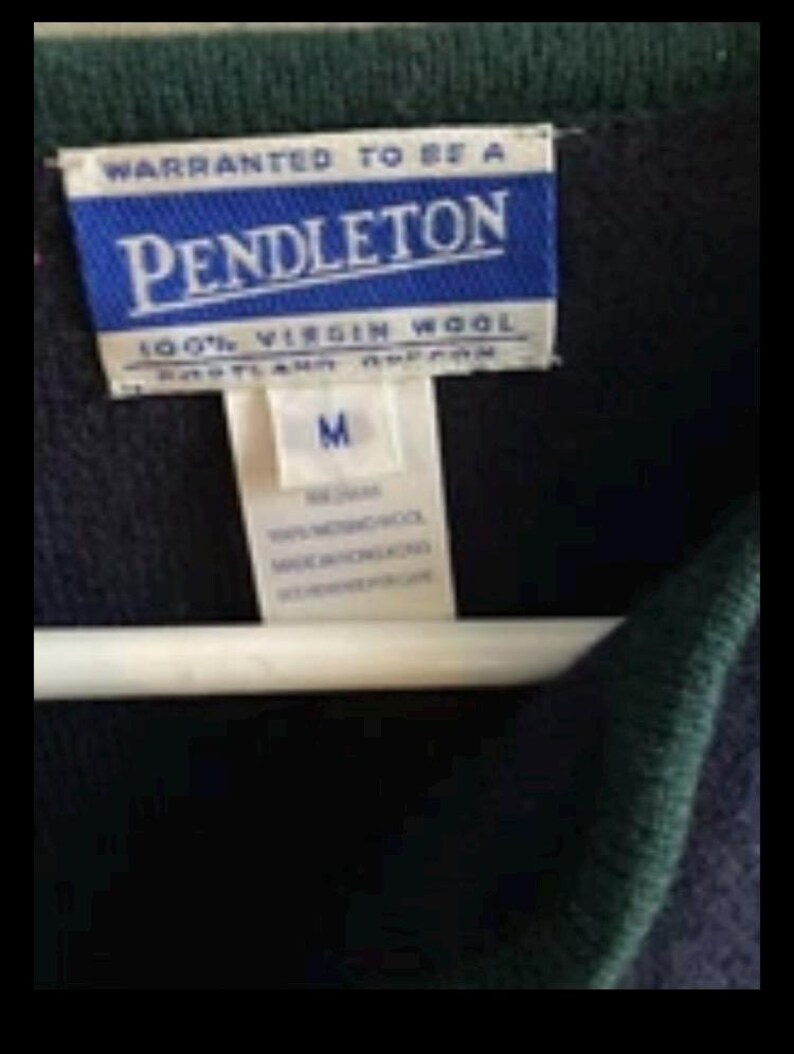 Puede incluir: Primer plano de un su&eacute;ter azul marino con ribete verde. La etiqueta dice "PENDLETON" en letras blancas sobre fondo azul, con "100% VIRGIN WOOL" debajo. Una etiqueta blanca indica la talla "M".