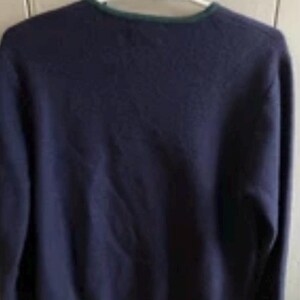 Puede incluir: Un su&eacute;ter azul marino con cuello redondo y mangas largas. El cuello y el dobladillo est&aacute;n ribeteados con una fina banda verde. El su&eacute;ter est&aacute; colgado de una percha blanca.