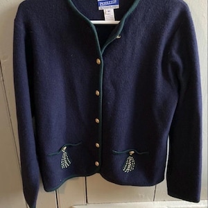 Könnte beinhalten: Marineblauer Cardigan mit grüner Paspelierung und goldenen Knöpfen. Der Cardigan hat zwei Taschen mit gestickten Quasten. Der Markenname "Pendleton" ist auf dem Etikett zu sehen. Der Cardigan hängt an einem weißen Kleiderbügel.
