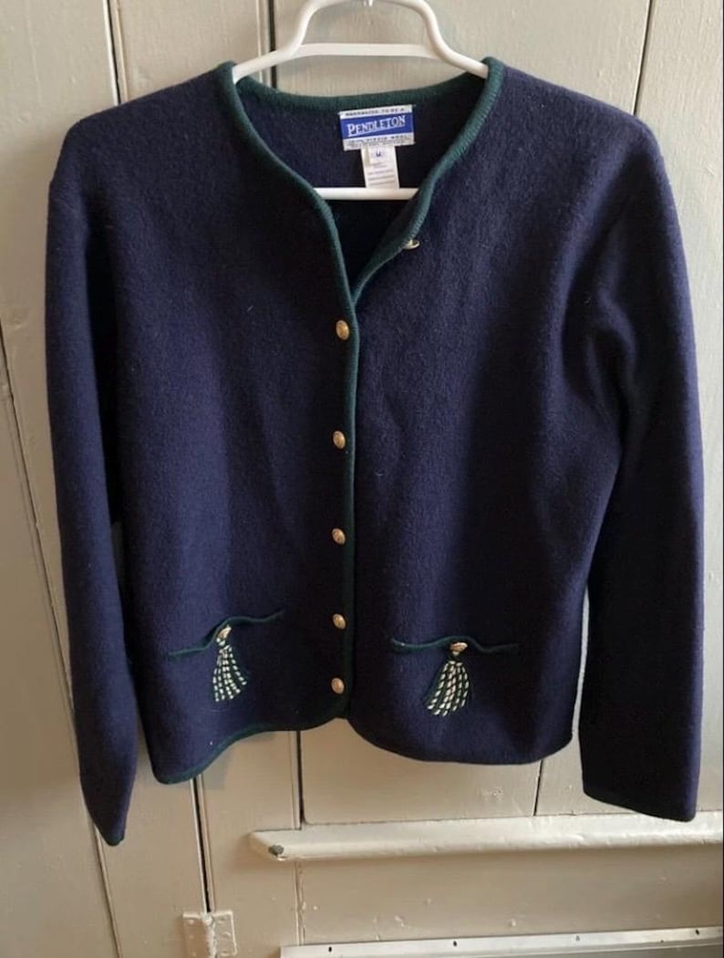 Puede incluir: C&aacute;rdigan azul marino con ribete verde y botones dorados. El c&aacute;rdigan tiene dos bolsillos con borlas bordadas. La marca "Pendleton" es visible en la etiqueta. El c&aacute;rdigan est&aacute; colgado de una percha blanca.