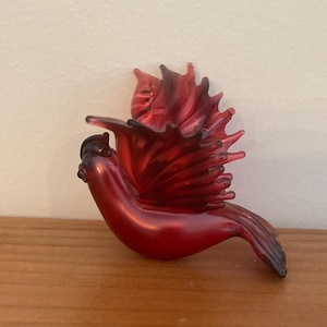 Könnte beinhalten: Rote Glas-Kardinal-Figur mit ausgebreiteten Flügeln. Der Vogel hat eine glänzende Oberfläche und sitzt auf einer Holzoberfläche. Die Flügel sind mit einem strukturierten Design versehen, und der Körper des Vogels ist ein sattes, leuchtendes Rot.