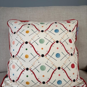 May include: Square cushion with a cream-coloured background featuring a repeating pattern of black lines, red wavy lines, and colourful circles. The cushion has a red border.