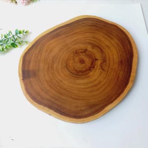 Puede incluir: Una tabla de cortar redonda de madera con borde natural. La tabla es de color marrón cálido con anillos marrones más oscuros, que muestran el patrón de crecimiento del árbol. La superficie es lisa y parece bien acabada, adecuada para la preparación de alimentos.
