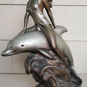 Op de afbeelding: Bronzen beeld van een vrouw die op een dolfijn rijdt. De vrouw draagt een vloeiende jurk en heeft lang haar. De dolfijn is zilverkleurig en komt uit een golfachtige basis.