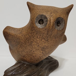 May include: A brown ceramic owl figurine with speckled brown paint and large, round eyes. The owl is perched on a brown, textured base.