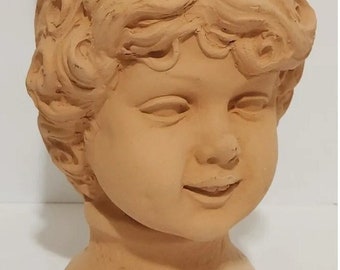 Op de afbeelding: Terracotta plantenbak in de vorm van een cherubijnhoofd met krullend haar. De plantenbak heeft een brede, open bovenkant en een voet.