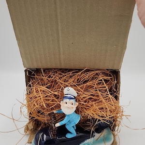 Puede incluir: Figura de temática náutica de un marinero dirigiendo un barco con forma de ballena. El marinero lleva gorro blanco y uniforme azul. La ballena es azul oscuro con base azul claro y blanca. En una caja de cartón con paja.