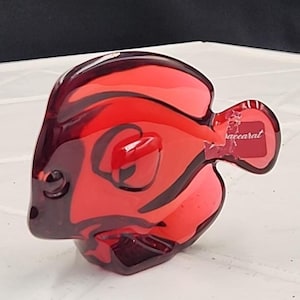 May include: A red glass fish ornament with a glossy finish. The fish has a detailed design with dark red accents and the word "baccarat" on the tail. The figurine is a decorative item.