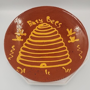 May include: A round, brown ceramic plate with a yellow beehive design in the center. The words "Busy Bees" are written above the beehive. The plate has a textured rim and is decorated with yellow bees and wavy lines.