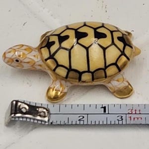 May include: A small, decorative turtle figurine. The turtle has a beige shell with a black geometric pattern. The head and legs are beige with gold accents. The figurine is approximately 2 inches long.