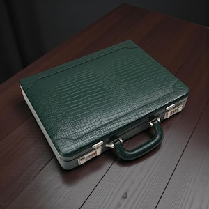 May include: A dark green briefcase with a textured, crocodile-like pattern. The briefcase has a handle, silver-colored clasps, and combination locks. The case is rectangular and sits on a dark wooden surface.