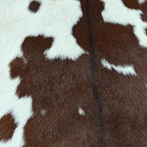 May include: Close-up of a cowhide rug with a natural pattern. The rug features a mix of white and brown patches, creating a unique and rustic aesthetic. The texture of the fur is visible.