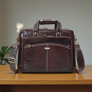 Puede incluir: Un maletín de cuero burdeos con asa superior y bandolera desmontable. La bolsa presenta un bolsillo frontal con cremallera, costuras blancas y herrajes dorados. El maletín está sobre una superficie de madera.