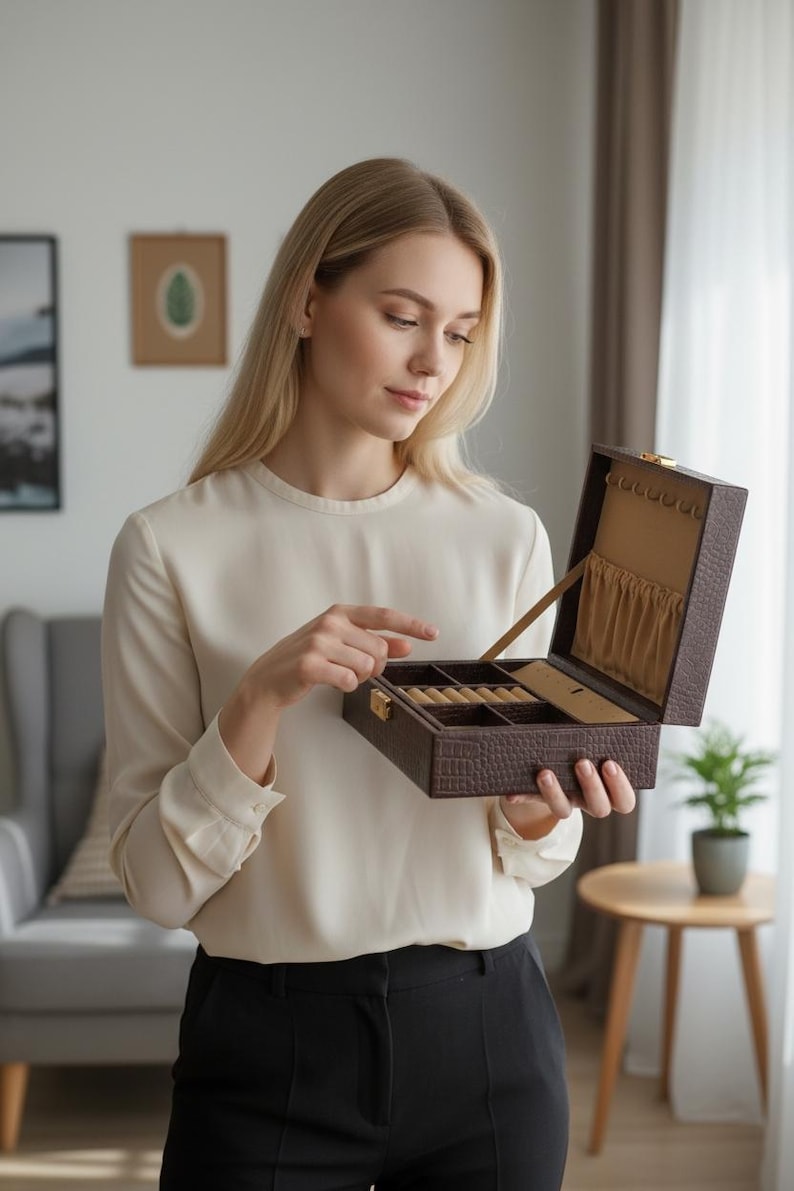 K&ouml;nnte beinhalten: Eine braune Schmuckschatulle mit Krokodilmuster ist ge&ouml;ffnet und zeigt F&auml;cher f&uuml;r Ringe und anderen Schmuck. Die Schatulle wird von einer Person gehalten, die eine cremefarbene Bluse und eine schwarze Hose tr&auml;gt. Der Innenraum ist mit beigem Stoff ausgekleidet.