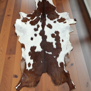 May include: A cowhide rug with a natural pattern of brown and white spots. The rug is irregularly shaped, with a soft, textured surface. The hide is laid on a hardwood floor.