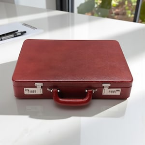 May include: A burgundy leather briefcase with a handle and combination locks. The rectangular case is closed and sits on a white surface. A pen and notepad are visible in the background.
