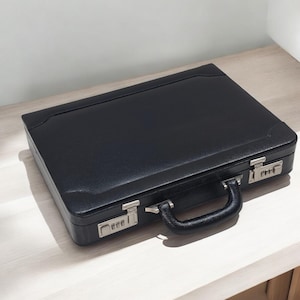 May include: A black leather briefcase with a combination lock closure and a black handle. The briefcase is rectangular and sits on a light-colored wooden surface. The hardware is silver-toned.