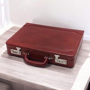 May include: A burgundy leather briefcase with silver-colored combination locks and a matching handle. The rectangular case is closed and sits on a light-colored surface. The briefcase has a classic, professional design.
