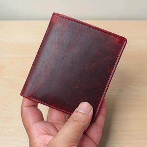 May include: A bifold wallet crafted from deep red leather with a distressed, matte finish. The wallet features visible tonal stitching along the edges and a rectangular shape with rounded corners, showcasing natural variations in the leather texture.