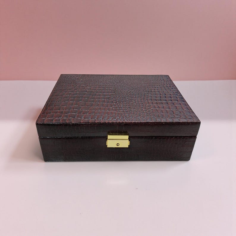 K&ouml;nnte beinhalten: Eine rechteckige, dunkelbraune Box mit einem strukturierten, krokodilartigen Muster. Die Box hat einen goldfarbenen Verschluss und Scharniere. Die Box wird wahrscheinlich zur Aufbewahrung oder Pr&auml;sentation von Schmuck oder anderen kleinen Gegenst&auml;nden verwendet.