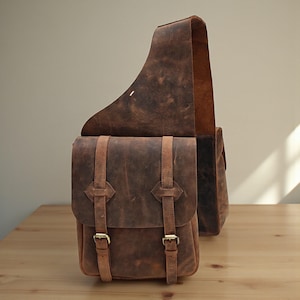 Puede incluir: Alforja de cuero marrón con bandolera. La bolsa tiene dos compartimentos con cierres de hebilla y está hecha de cuero envejecido. La bolsa está sobre una superficie de madera.