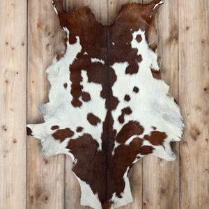 May include: A cowhide rug with a brown and white spotted pattern. The rug is laid out on a light brown wooden surface. The hide has a natural shape with irregular edges, showcasing the animal's original form.