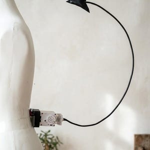 May include: A mechanical, motorized black tail accessory attached to a white mannequin torso. The device features a white plastic motor housing with a circular speaker grille, connected to a long, thin, flexible black wire ending in a pointed tip.