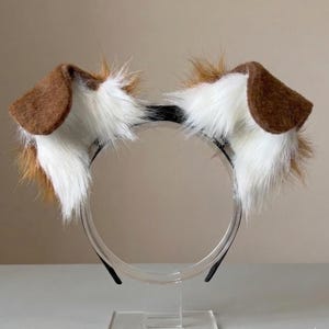 May include: A headband featuring two plush animal ears made of long, white and brown faux fur. The ears are positioned symmetrically on a thin, black plastic band. The fur texture is shaggy and layered, with brown tips and white inner sections.