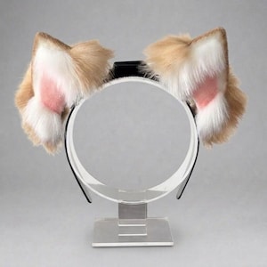 Może przedstawiać: Opaska do włosów z pluszowymi, futrzanymi uszami kota. Uszy są w kolorze brązowym z białymi i różowymi akcentami, przymocowane do czarno-białej opaski. Prezentowane na przezroczystym, akrylowym stojaku.