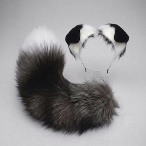 May include: A set consisting of a fluffy, long, curved faux fur tail in shades of black, grey, and white, paired with a matching headband featuring two rounded, upright animal ears that are black at the tips and white at the base.