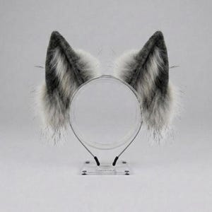 May include: A pair of faux fur animal ears in shades of grey, white, and black, mounted on a clear acrylic headband display stand. The ears feature long, textured synthetic fibres with a gradient colour pattern and pointed, triangular shapes.