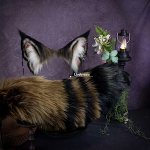 Peut inclure: Un ensemble d'oreilles d'animal en fausse fourrure avec une queue rayée assortie. Les oreilles sont brunes, noires et blanches avec des chaînes argentées et des breloques rouges. Le texte "Lively ears" est visible. La queue présente des bandes alternées de fourrure brune et noire.