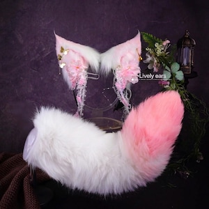 Può includere: Un set di orecchie da animale in pelliccia sintetica bianca e rosa con coda abbinata. Le orecchie sono decorate con fiori in tessuto rosa, ciondoli a farfalla dorati e perline di cristallo pendenti. Il testo "Lively ears" è stampato.