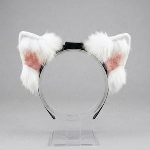 Op de afbeelding: Een zwarte haarband met twee pluizige, witte kattenoren. De binnenkant van elk oor is roze. De oren zijn gemaakt van zacht imitatiebont. De haarband wordt op een transparante standaard gepresenteerd.