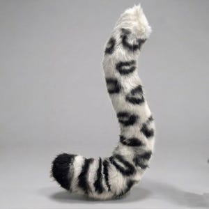 May include: A long, curved faux fur tail featuring a white base color with black leopard-style spots and a solid black tip. The plush, textured material is arranged in a vertical, slightly S-shaped posture, showcasing a dense, soft appearance.