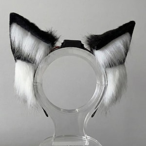 Può includere: Una fascia per capelli con orecchie da volpe in finta pelliccia. Le orecchie sono nere e bianche, con una consistenza soffice. La fascia è esposta su un supporto di plastica trasparente.