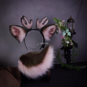 Puede incluir: Una diadema con dos orejas de animal puntiagudas de peluche con pelo blanco y marrón, acompañadas de dos pequeñas astas lisas de color claro. Debajo de la diadema hay una cola grande y esponjosa de piel sintética bicolor.