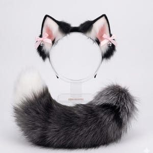 Può includere: Un set cosplay con cerchietto con orecchie da gatto nere e bianche, dettaglio interno rosa e piccoli fiocchi rosa con campanellini argentati. Una coda soffice grigia e bianca abbinata completa il set. Gli articoli sono esposti su un supporto trasparente.