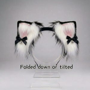 Puede incluir: Una diadema con dos orejas de gato de piel sintética en blanco con puntas negras y detalles interiores rosas. Cada oreja está adornada con un pequeño lazo negro y un cascabel plateado. El texto "Folded down or tilted" aparece debajo.
