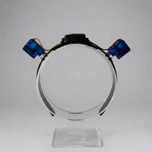 Puede incluir: Una diadema circular negra con dos pequeños servomotores electrónicos azules sujetos a los lados. Los motores están conectados mediante cables finos de color naranja y negro a un componente electrónico central negro montado en la diadema.