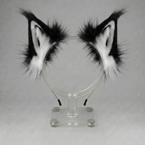 May include: A pair of pointed animal ears attached to a thin headband. The ears are covered in long, fluffy faux fur with a black outer edge and a white inner section. The headband is made of a thin, flexible material, designed to be worn on the head.