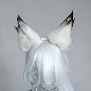 May include: A pair of tall, pointed animal ears made of white faux fur with dark grey to black tips. The ears are attached to a white headband and are positioned atop a head of long, straight, white hair that covers the forehead and sides.