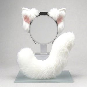 Op de afbeelding: Een witte set van een kattenstaart en hoofdband van imitatiebont. De hoofdband heeft twee witte kattenoren met roze binnenkant. De staart is gebogen en pluizig. De set wordt gepresenteerd op een transparante standaard tegen een witte achtergrond.