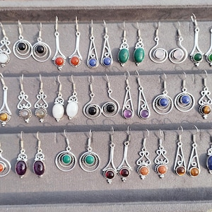 May include: A selection of silver earrings featuring diverse gemstones like red, blue, green, and black. The earrings are presented on a grey tray, displaying a range of designs and shapes, suitable for different styles.