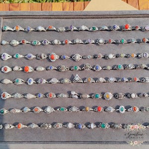 Peut inclure: Une présentation de nombreuses bagues en argent avec divers sertissages de pierres précieuses, notamment des pierres rouges, bleues, vertes et blanches. Les bagues sont disposées avec soin sur une surface grise, présentant une variété de modèles et de tailles. Les bagues sont de Gemstone Grace Co.