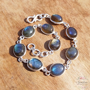 Op de afbeelding: Een zilveren armband met ovale labradoriet edelstenen. De stenen vertonen iriserende blauwe en groene tinten, gezet in een schakelketting met een sluiting. De armband wordt gepresenteerd op een houten oppervlak.