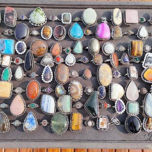 Puede incluir: Una colección de anillos de piedras preciosas expuestos sobre una superficie gris. Los anillos presentan diversas formas y colores, incluyendo turquesa, ojo de tigre y piedras rosas, engastados en metal plateado. Los anillos están dispuestos en filas, mostrando una variedad de diseños.