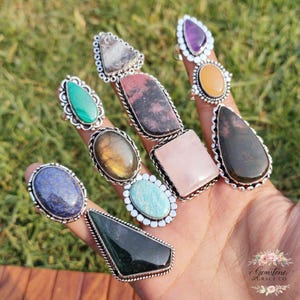 Könnte beinhalten: Eine Sammlung von Edelsteinringen in verschiedenen Formen und Farben, darunter grüner Malachit, violetter Amethyst und rosa Rosenquarz. Die Ringe sind in silberfarbenem Metall gefasst und auf einer Hand vor unscharfem grünem Hintergrund präsentiert.