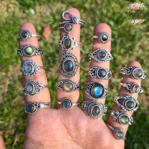 Könnte beinhalten: Eine Sammlung von Silberringen mit verschiedenen Labradorit-Edelsteinen. Die Ringe weisen unterschiedliche Designs auf, darunter ovale, runde und verzierte Fassungen. Die Edelsteine zeigen eine Reihe von Farben, darunter Blau, Grün und Grau. Die Ringe werden auf Fingern präsentiert.