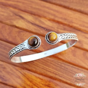 Puede incluir: Una pulsera de plata con dos piedras preciosas redondas de ojo de tigre. La pulsera tiene un diseño abierto detallado con un borde trenzado alrededor de las piedras preciosas y patrones en forma de hojas. Las piedras preciosas son de color marrón cálido.